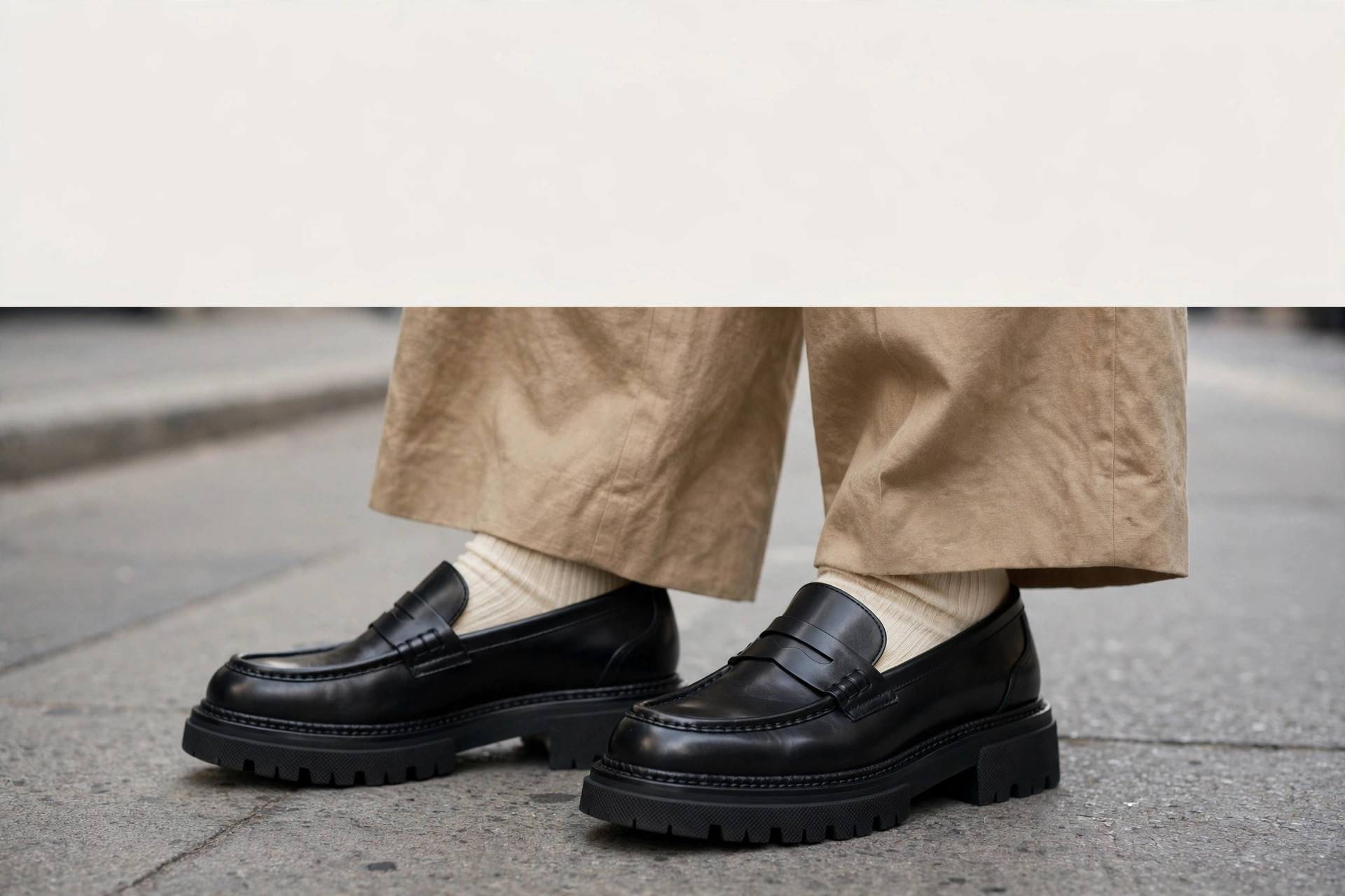 Chunky black loafers worn with white socks and wide trousers on a city street
